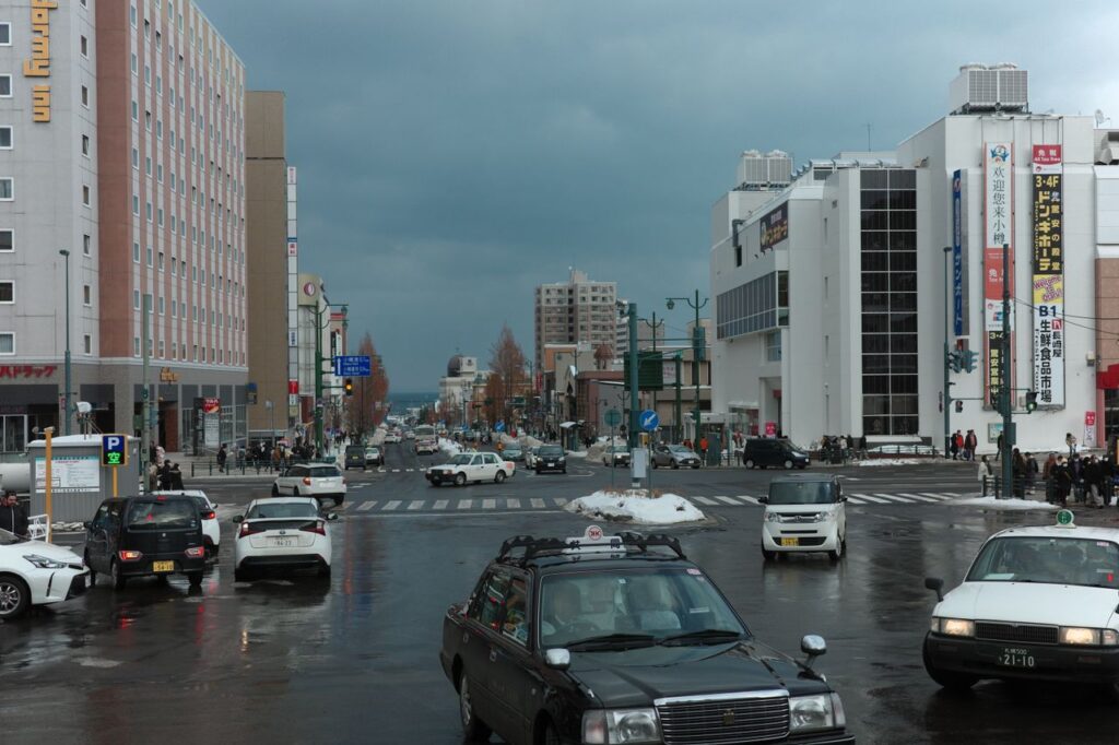 A busy winter street intersection in Otaru, Japan with cars and pedestrians on a cloudy day.