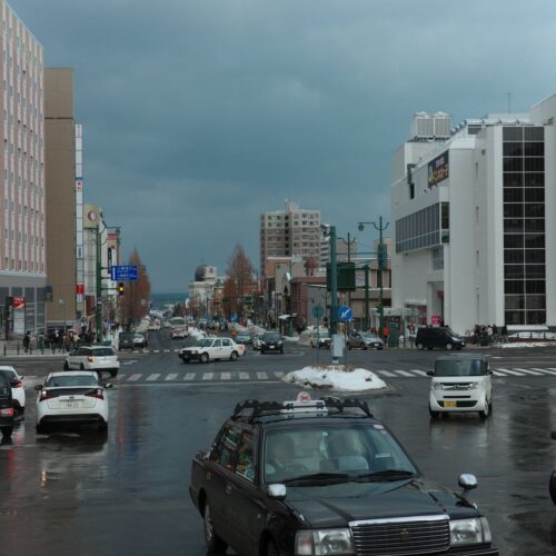 A busy winter street intersection in Otaru, Japan with cars and pedestrians on a cloudy day.