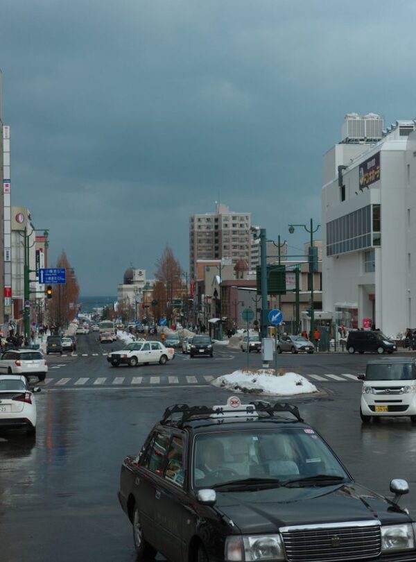 A busy winter street intersection in Otaru, Japan with cars and pedestrians on a cloudy day.