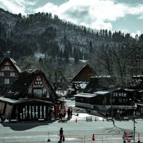 From above of old wooden houses near asphalt road and mountains with growing trees and unrecognizable people walking in village under sky with clouds
